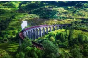 Gőzmozdony vörös vagonokkal a Glenfinnan-viadukton, körülötte zöld dombok