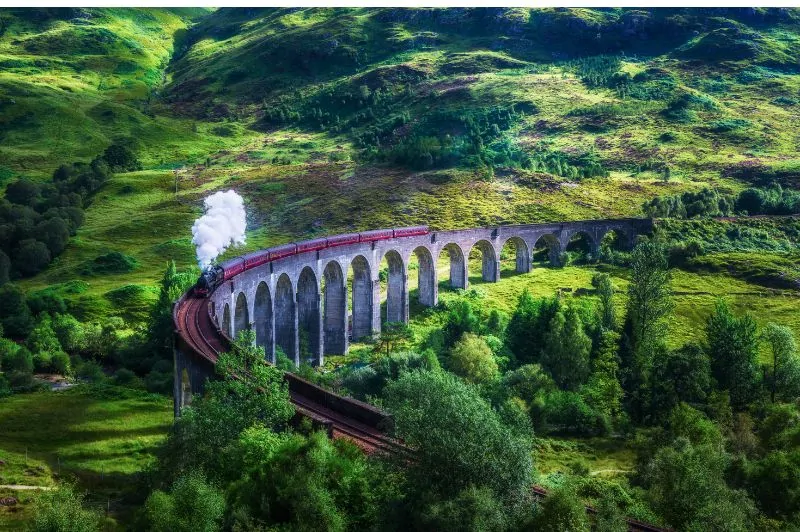 Gőzmozdony vörös vagonokkal a Glenfinnan-viadukton, körülötte zöld dombok