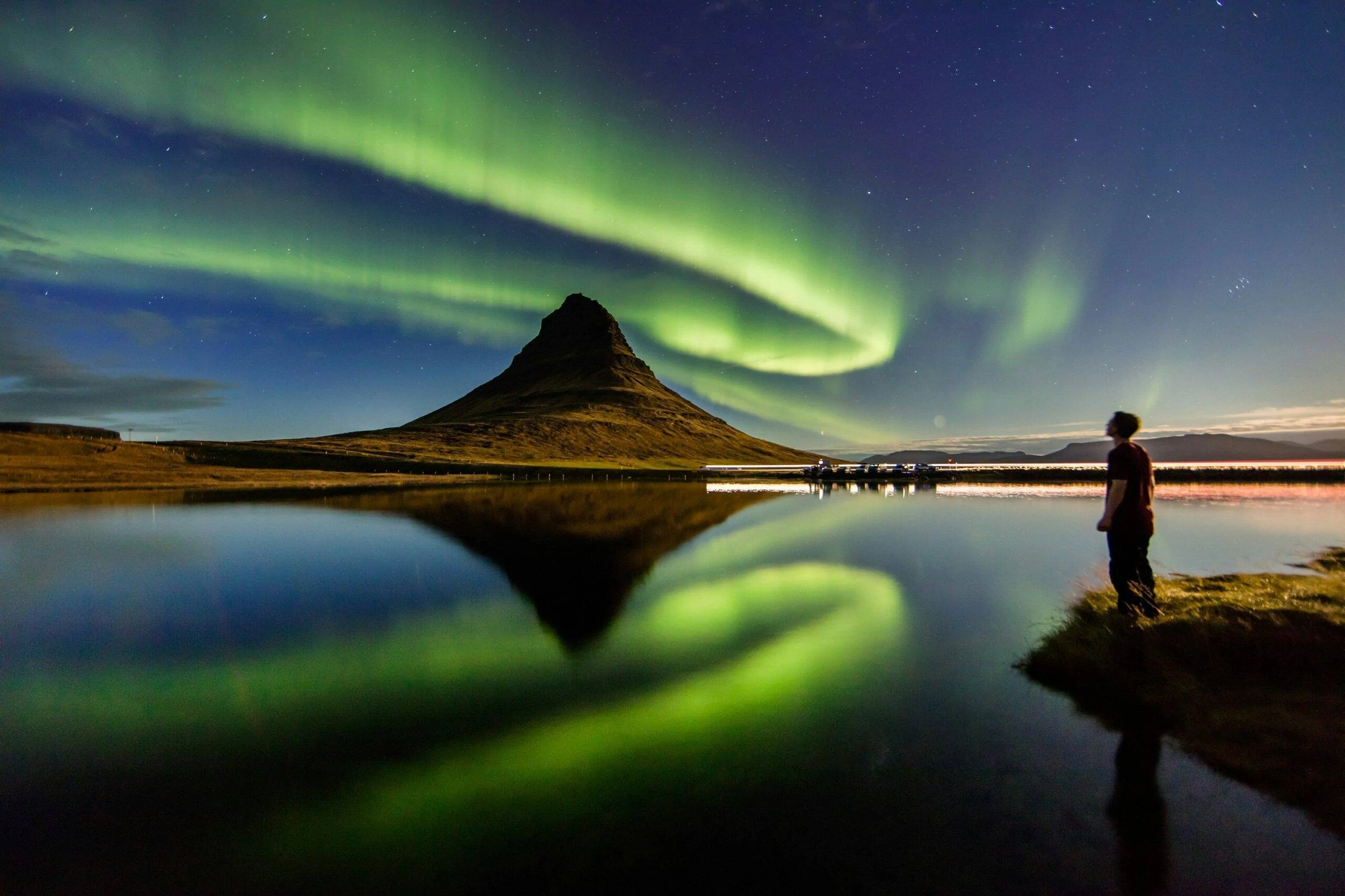 Zöld sarki fény a Kirkjufell hegy felett, vízparton álló alakkal